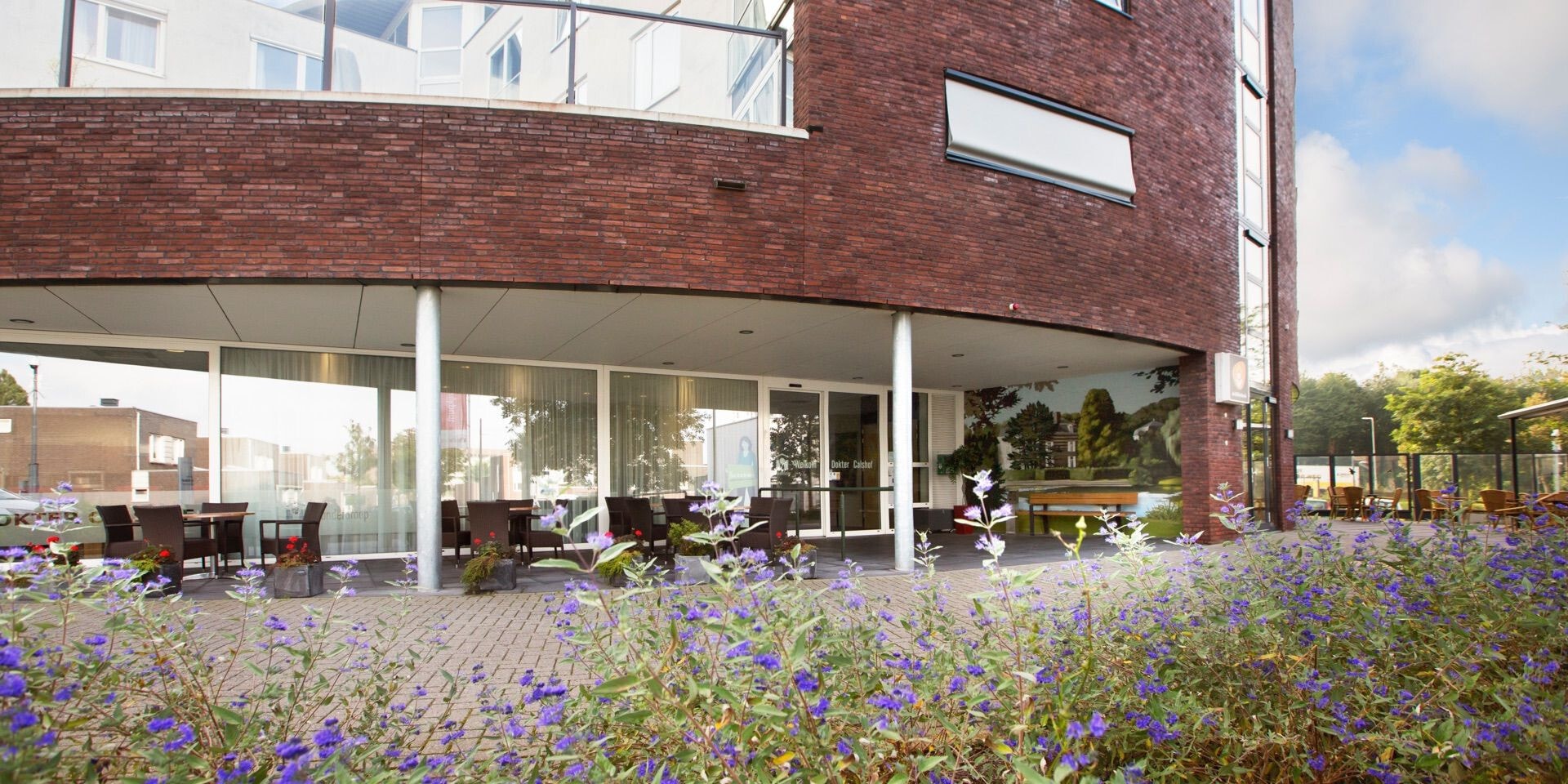 De entree van een modern bakstenen wooncomplex met een terras onder de overkapping, omgeven door een verzorgde tuin met paarse bloemen op de voorgrond.