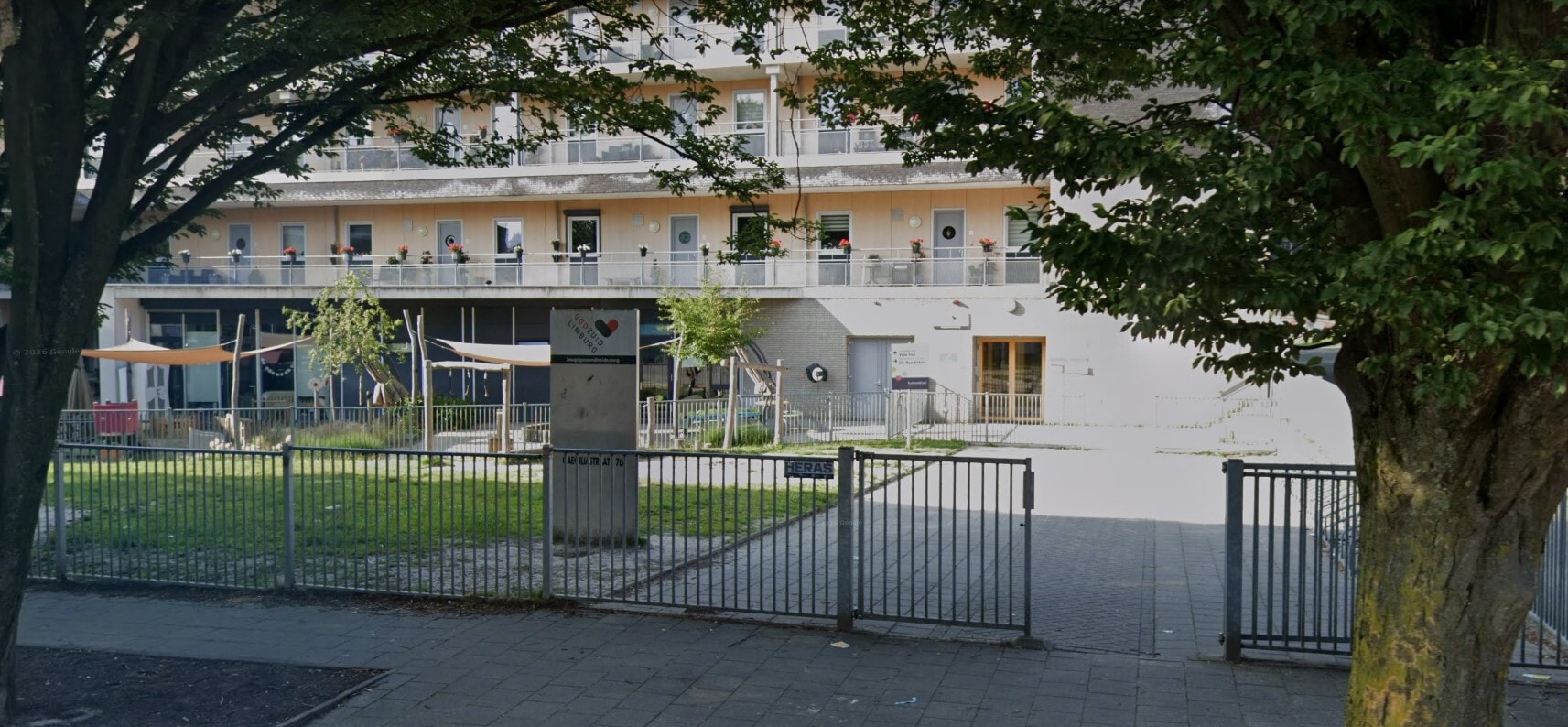 Een modern appartementencomplex met galerijen vol bloembakken, gelegen achter een omheind terrein met een gazon en schaduwdoeken.