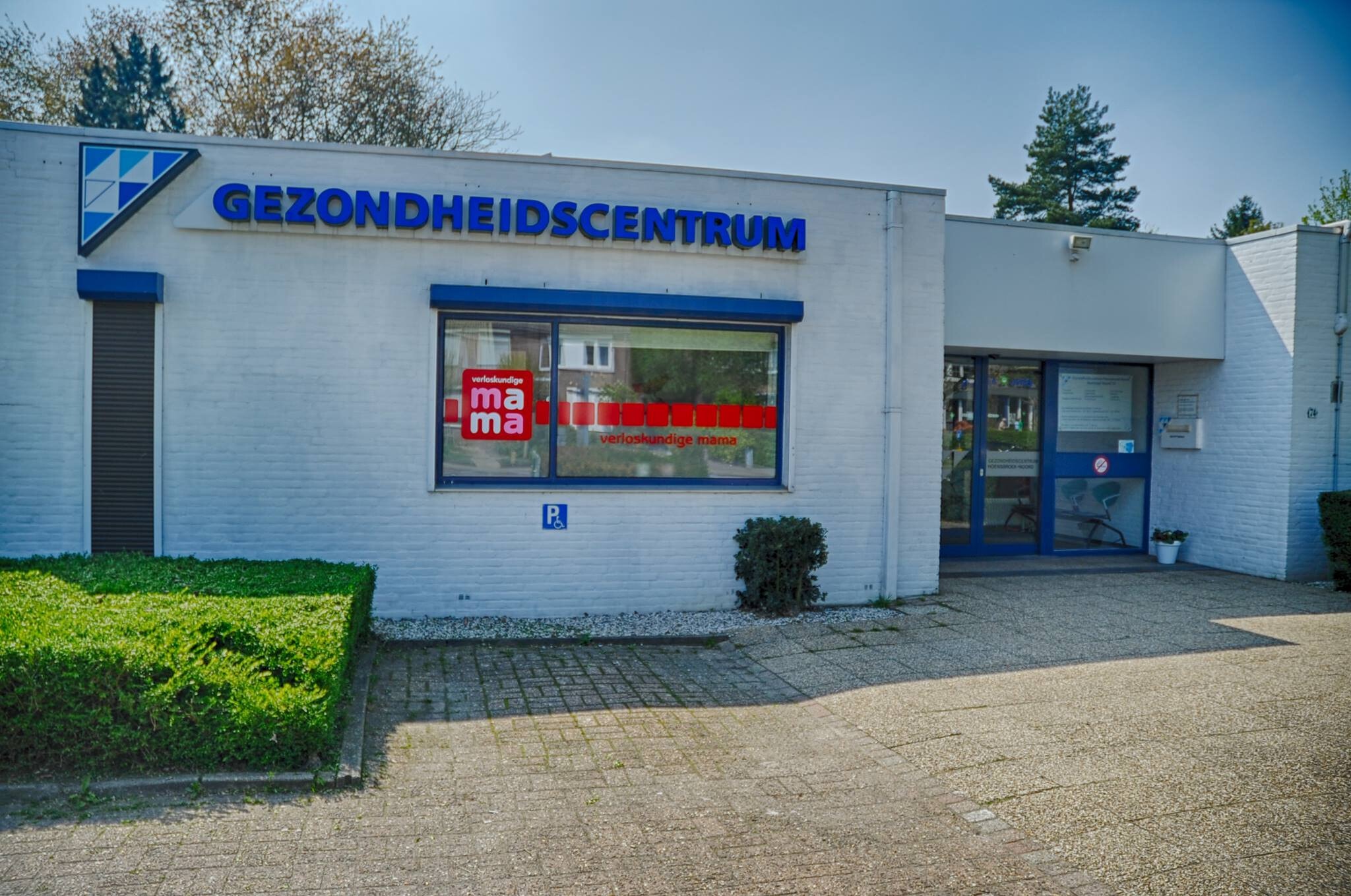 De witte gevel van een gezondheidscentrum met blauwe letters, waarin ook een verloskundigenpraktijk is gevestigd, gefotografeerd op een zonnige dag.