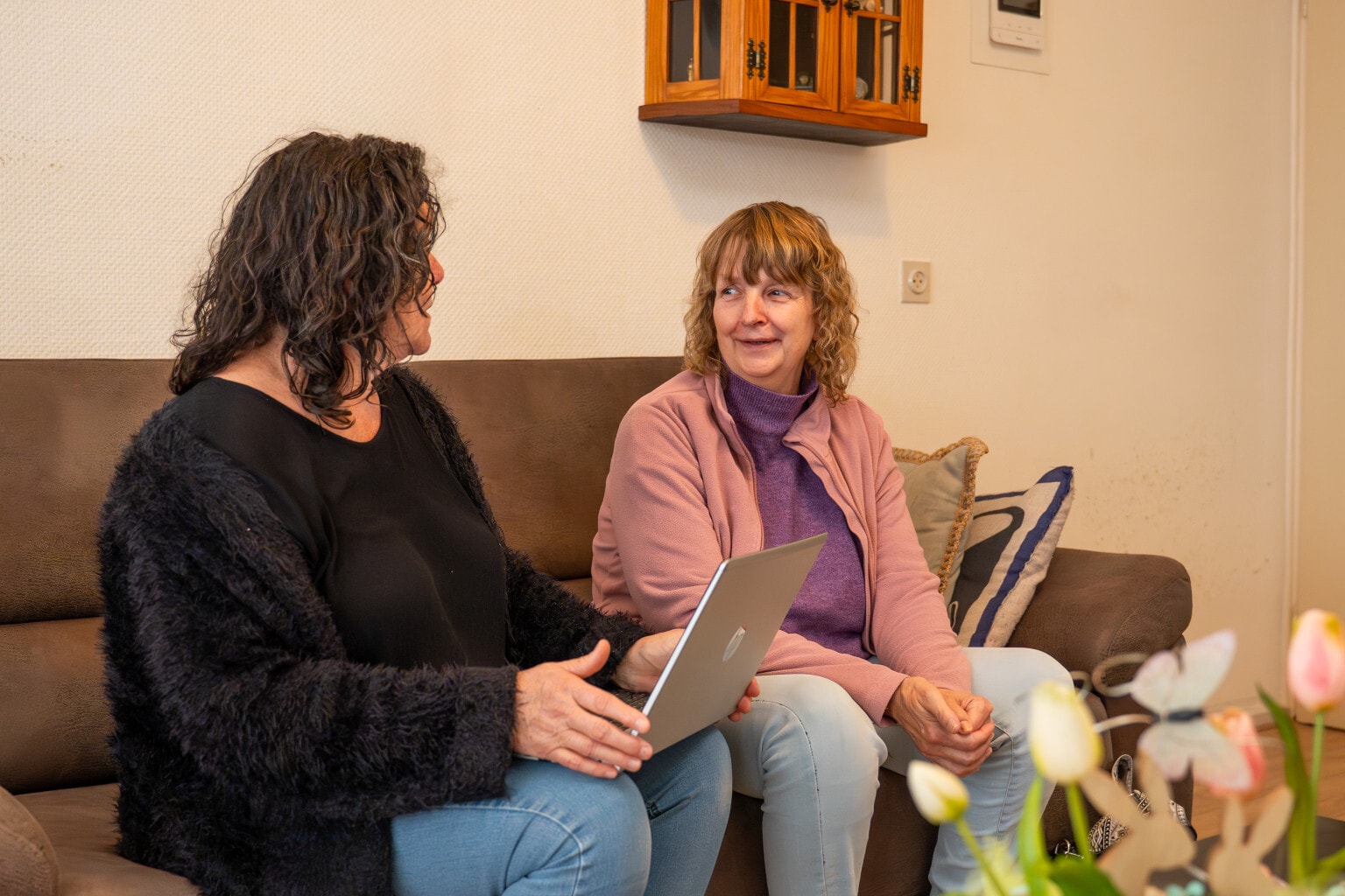Twee vrouwen zitten naast elkaar op een bruine bank. Een vrouw heeft een opengeklapte laptop op haar schoot.