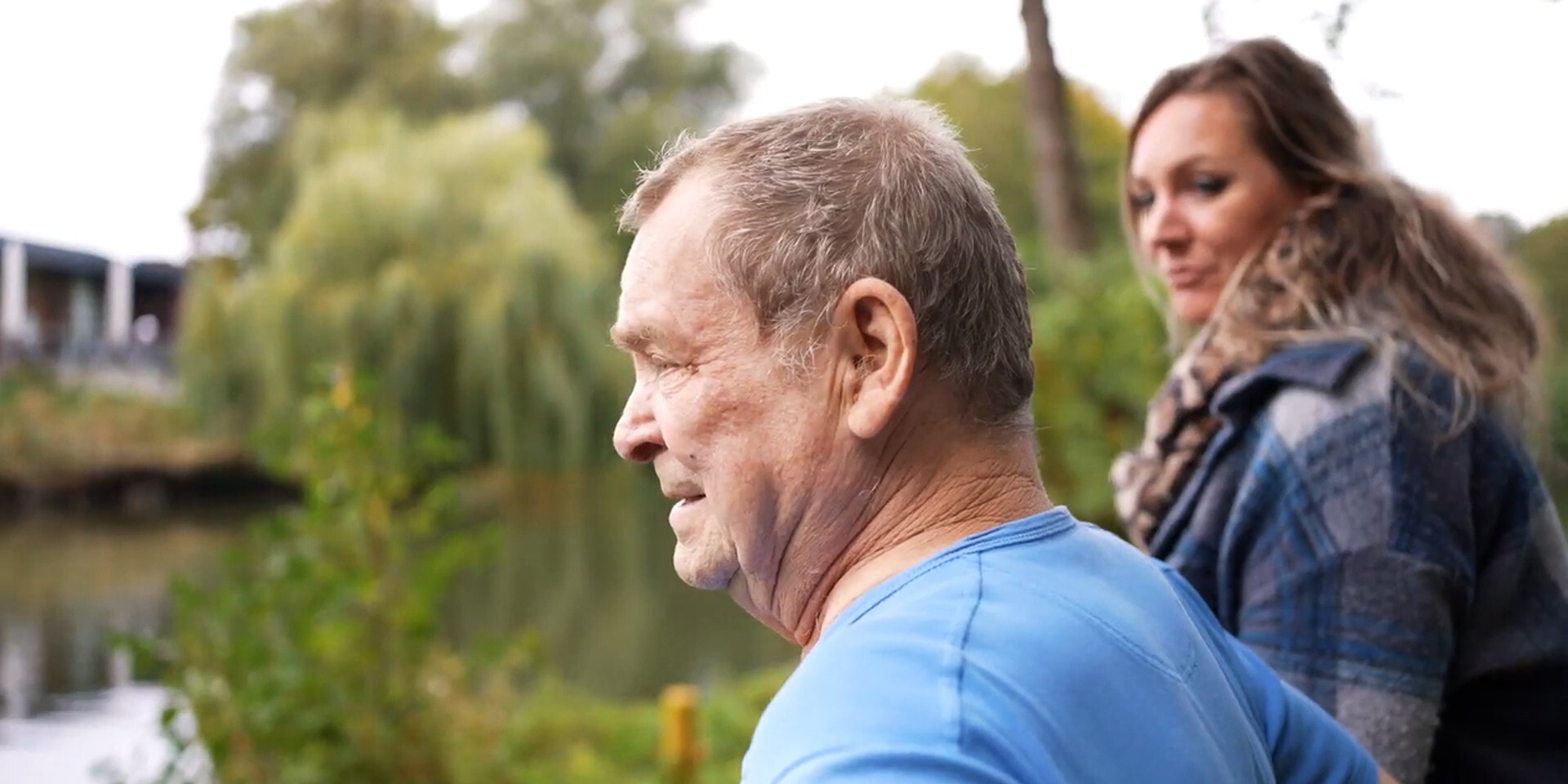 Een zijaanzicht van een oudere man in een blauw T-shirt die voor zich uit kijkt, naast hem zit een vrouw. Ze bevinden zich buiten in een parkachtige omgeving met een vijver en bomen.