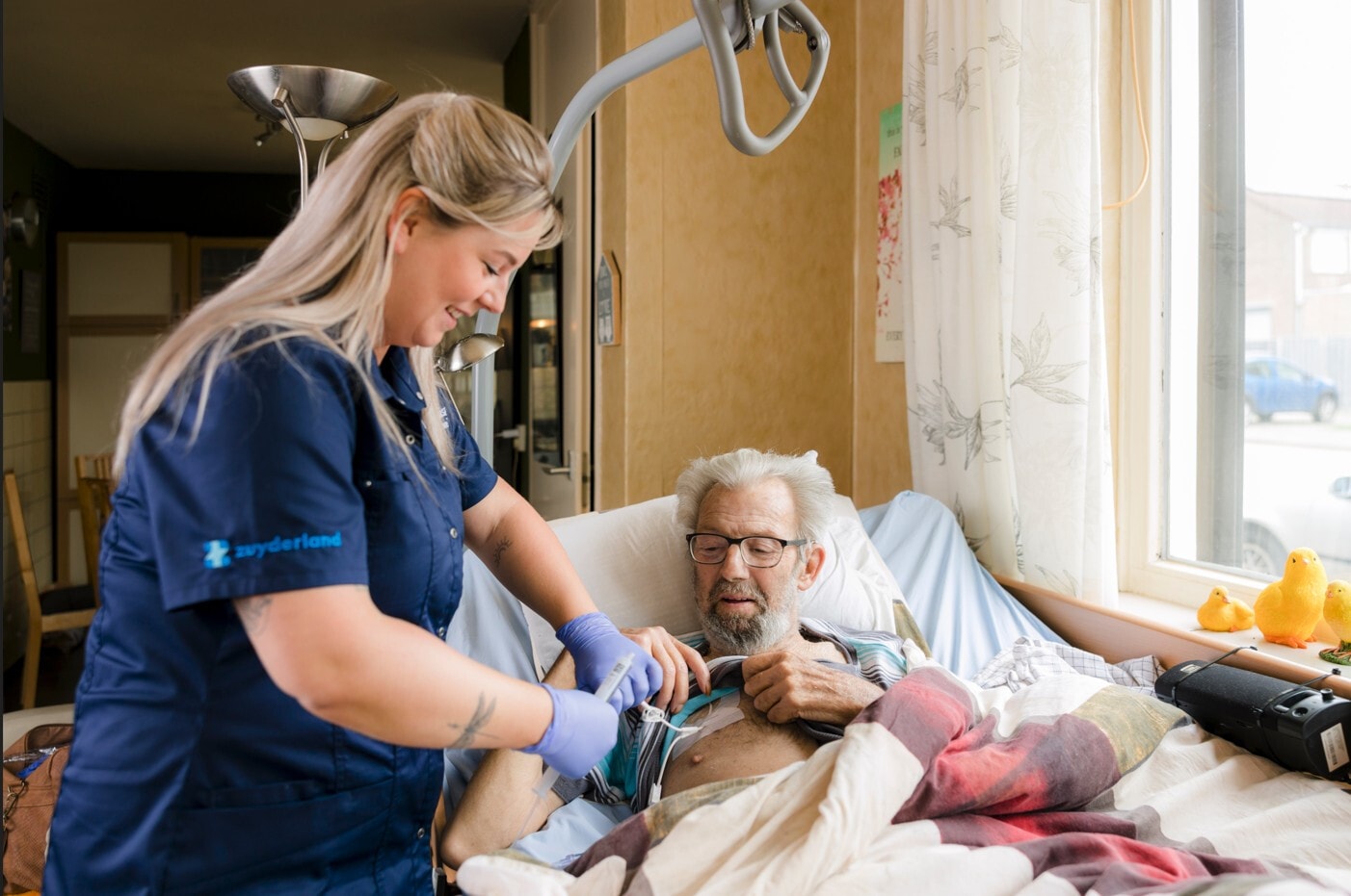Een zorgverlener van Zuyderland voert met een glimlach een medische handeling uit bij een oudere man die in een zorgbed ligt.