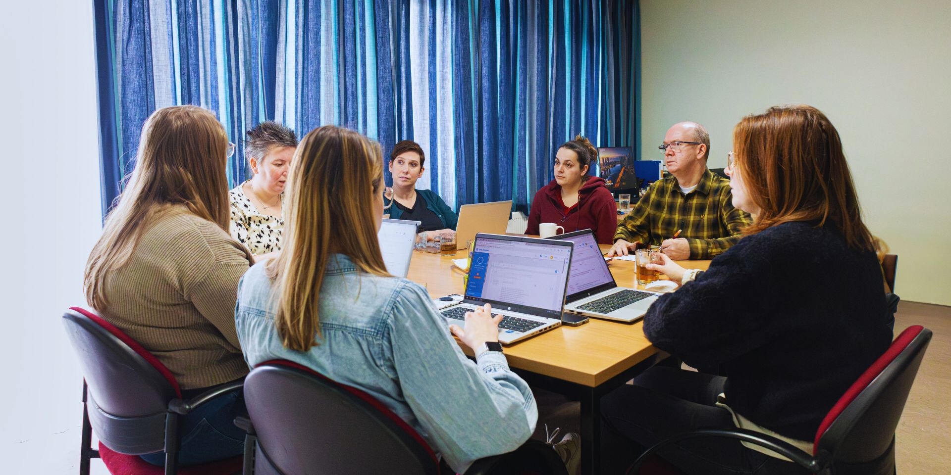 Een groep collega's zit rond een tafel voor een vergadering of overleg, waarbij enkelen gebruikmaken van laptops in een kantoorruimte met blauwe gordijnen.