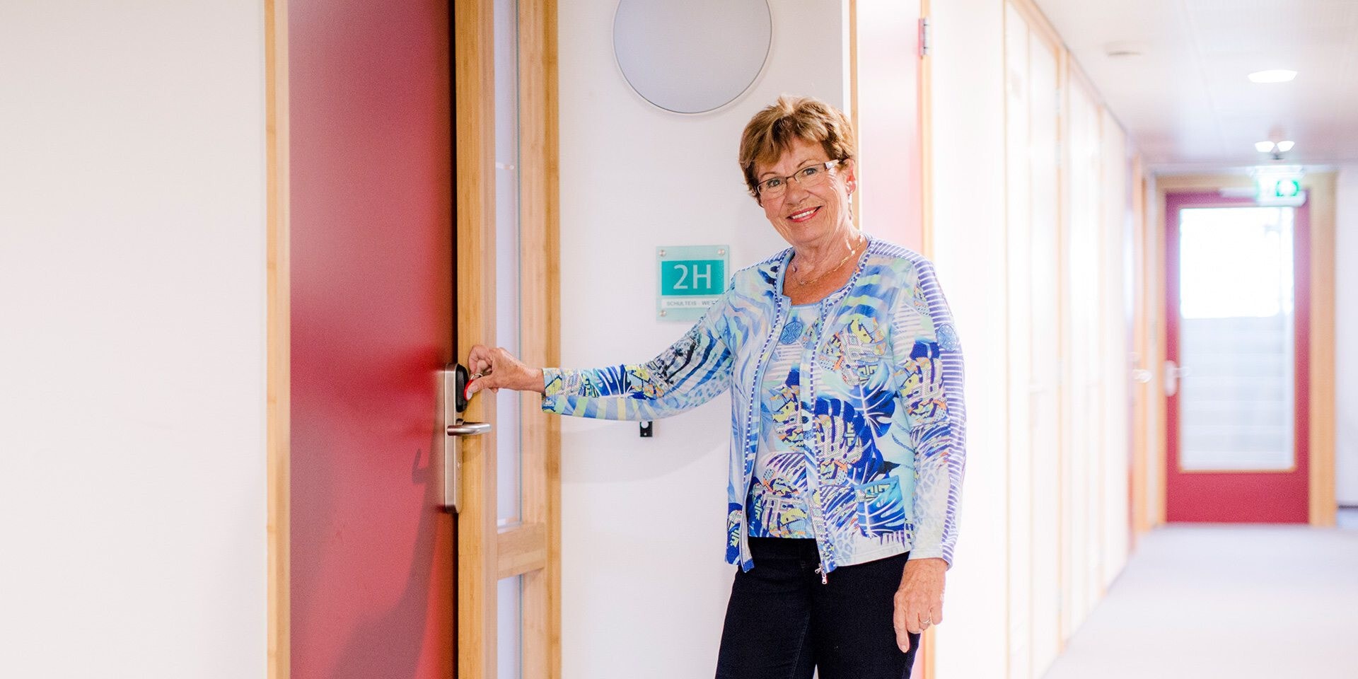Een glimlachende vrouw staat in een gang van een gebouw en opent een rode deur met een elektronische sleutel.