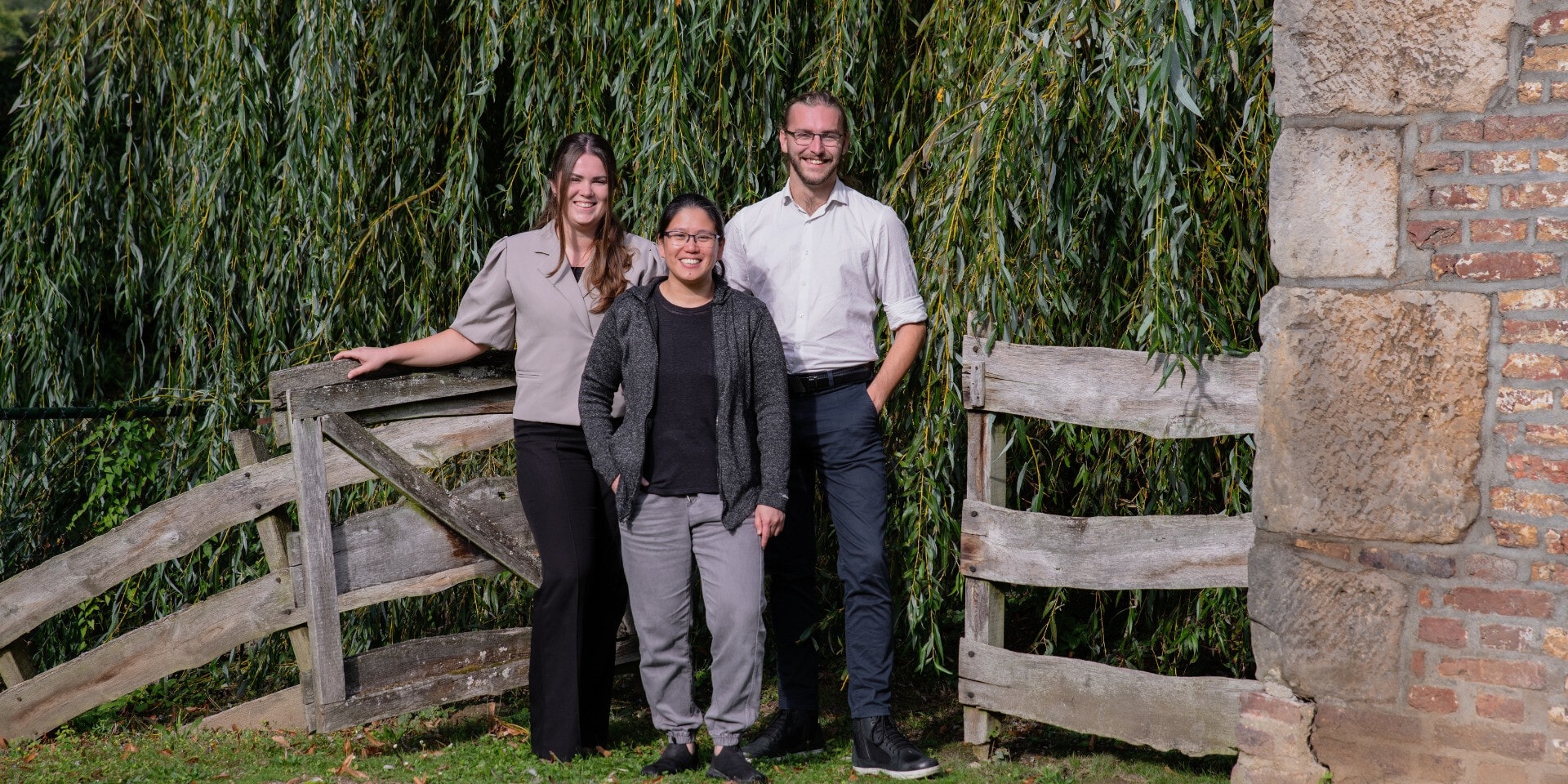 Twee lachende vrouwen en man staan voor een houten hek aan het water.