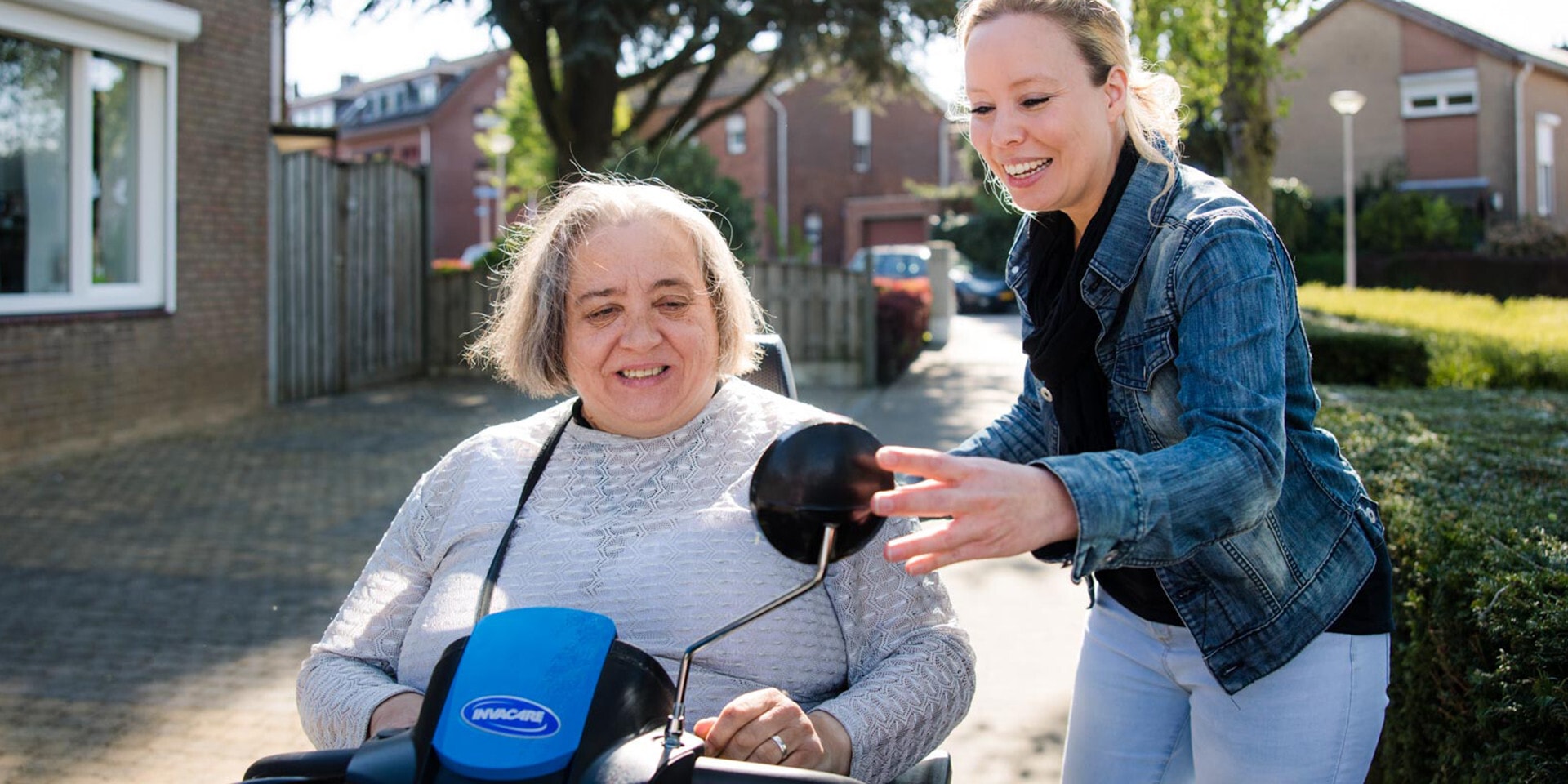 Jonge vrouw loopt naast een oudere vrouw in een scootmobiel en corrigeert haar spiegel.