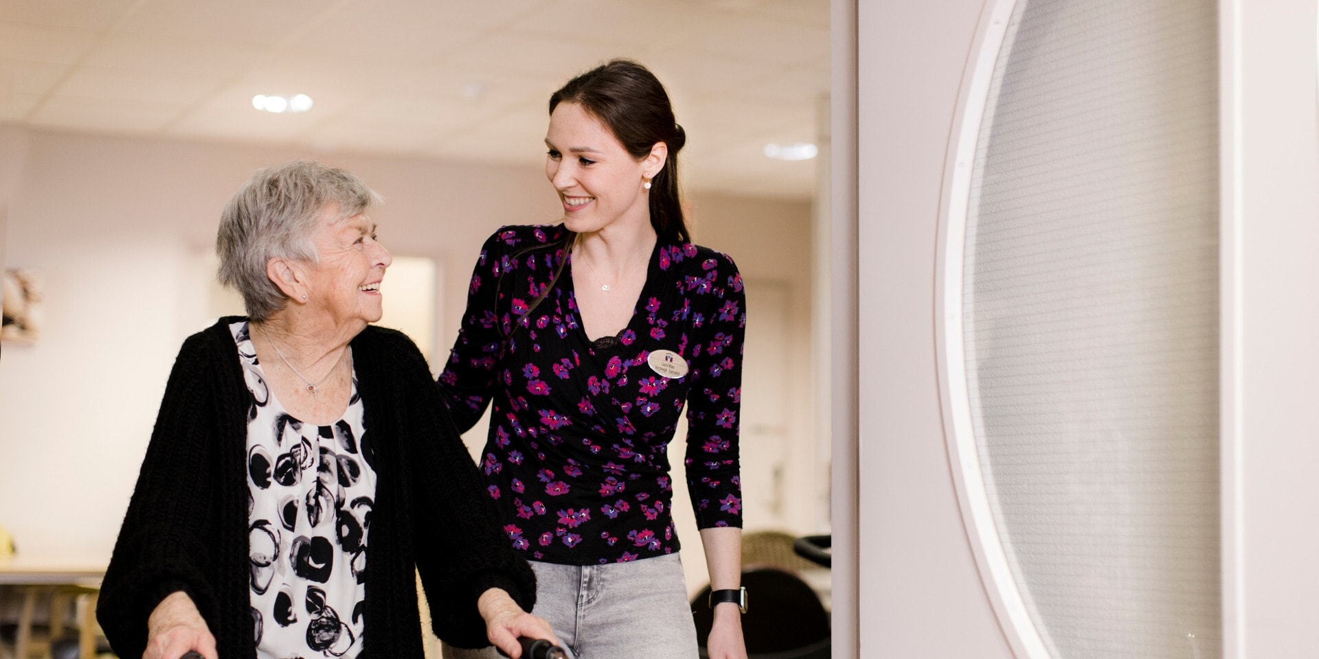 Oudere vrouw achter een rollator loopt naast een jonge vrouwelijke medewerkster van MeanderGroep.