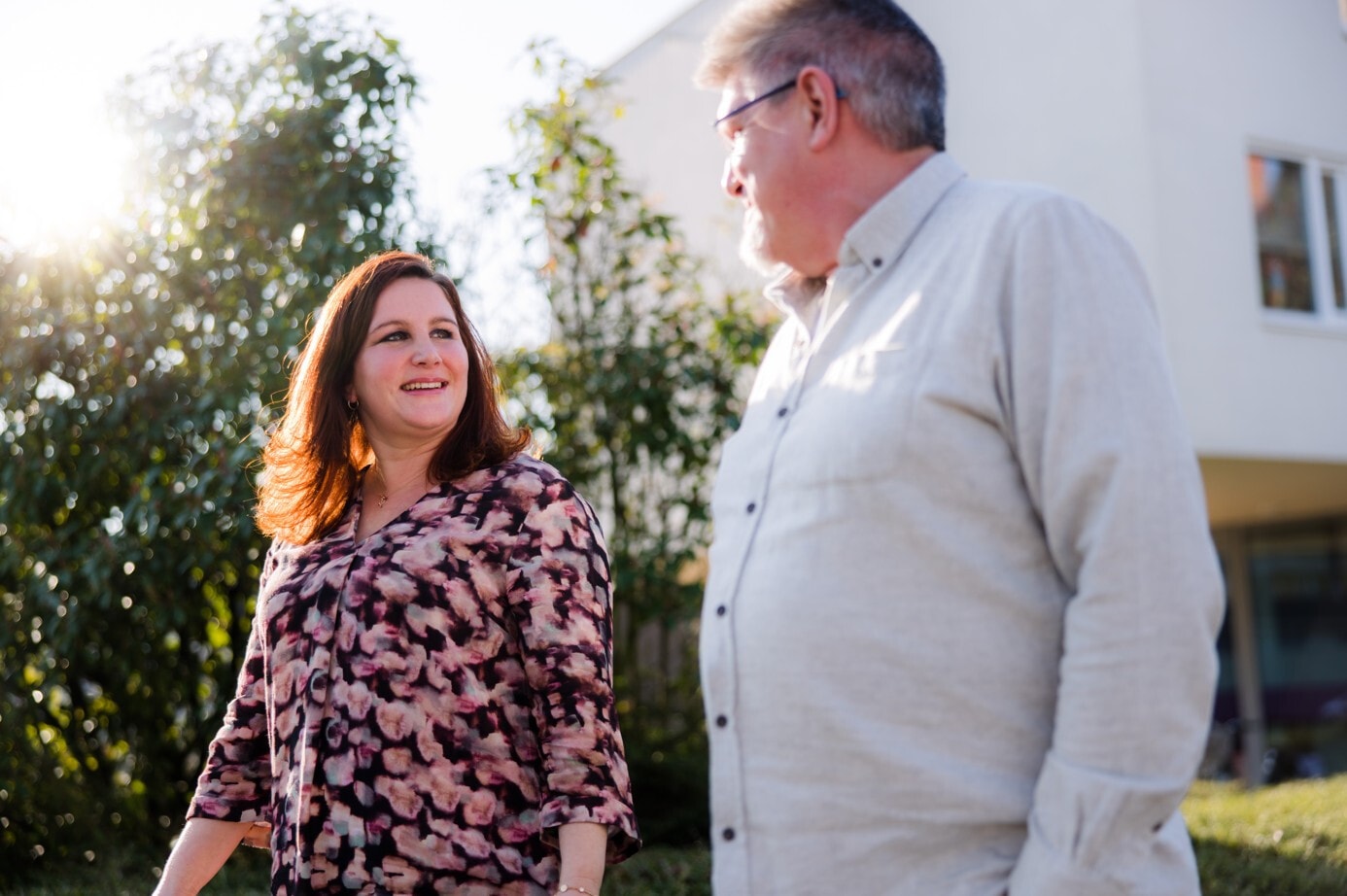 Een glimlachende vrouw loopt buiten en kijkt op naar een man die naast haar staat. De zon schijnt door de bladeren van een boom op de achtergrond.