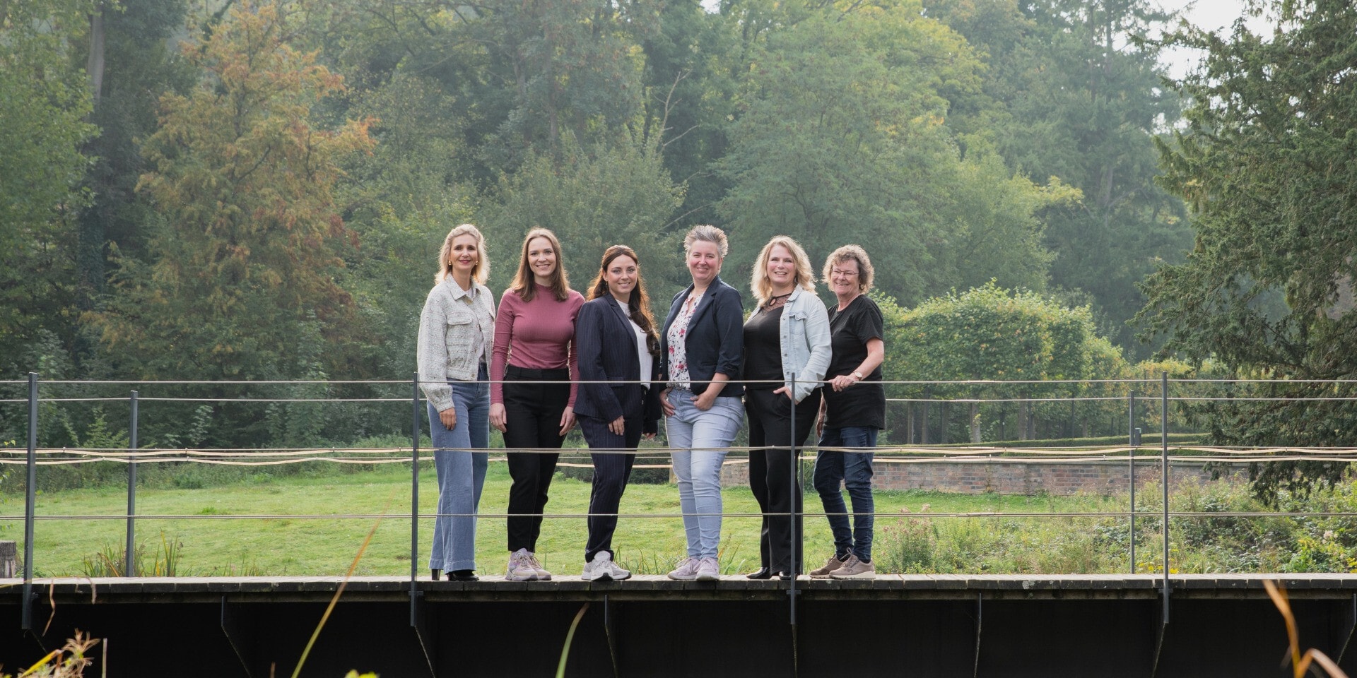 Zes vrouwen staan glimlachend naast elkaar op een houten brug.