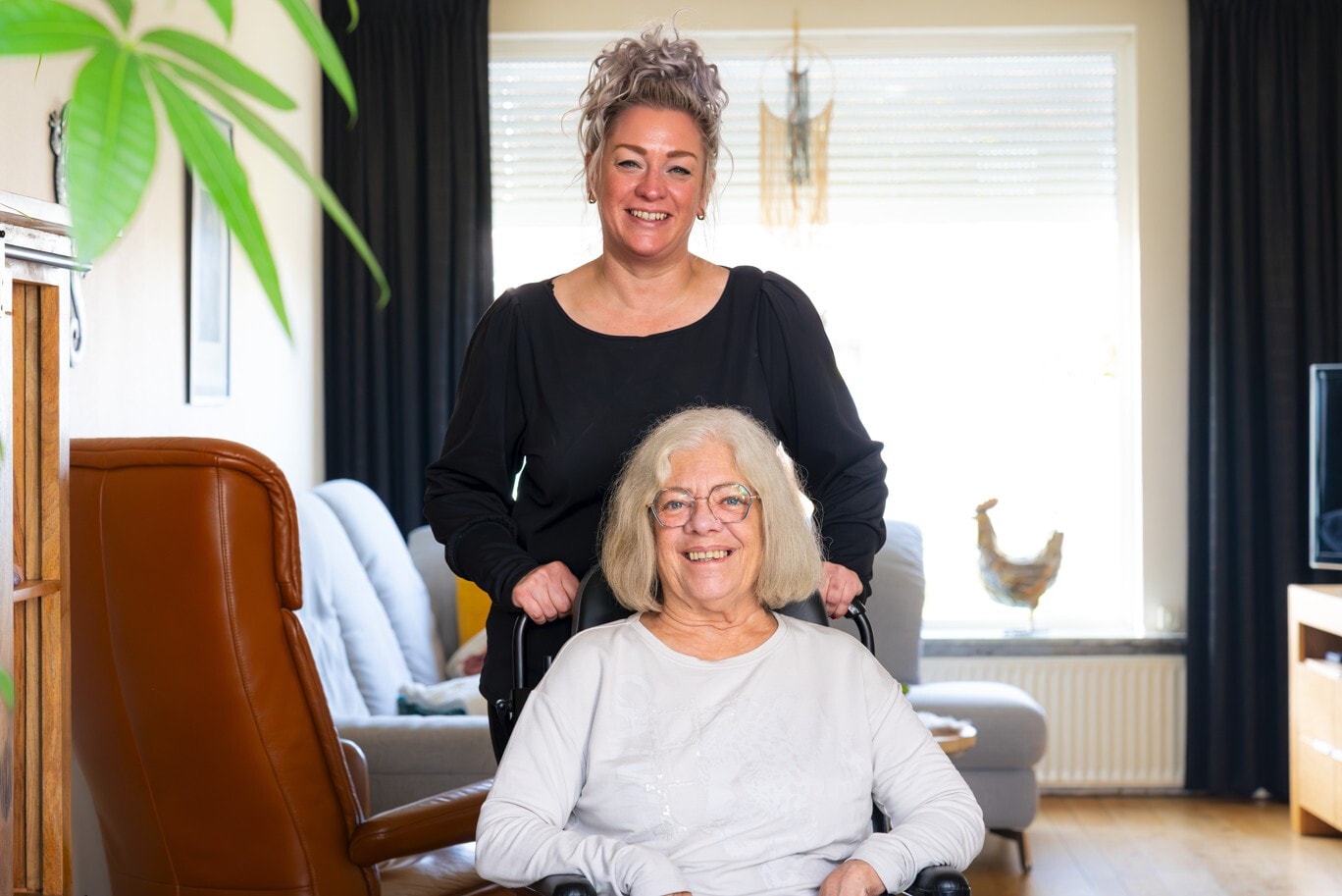 Een glimlachende vrouw staat achter een oudere vrouw in een rolstoel in een lichte woonkamer.