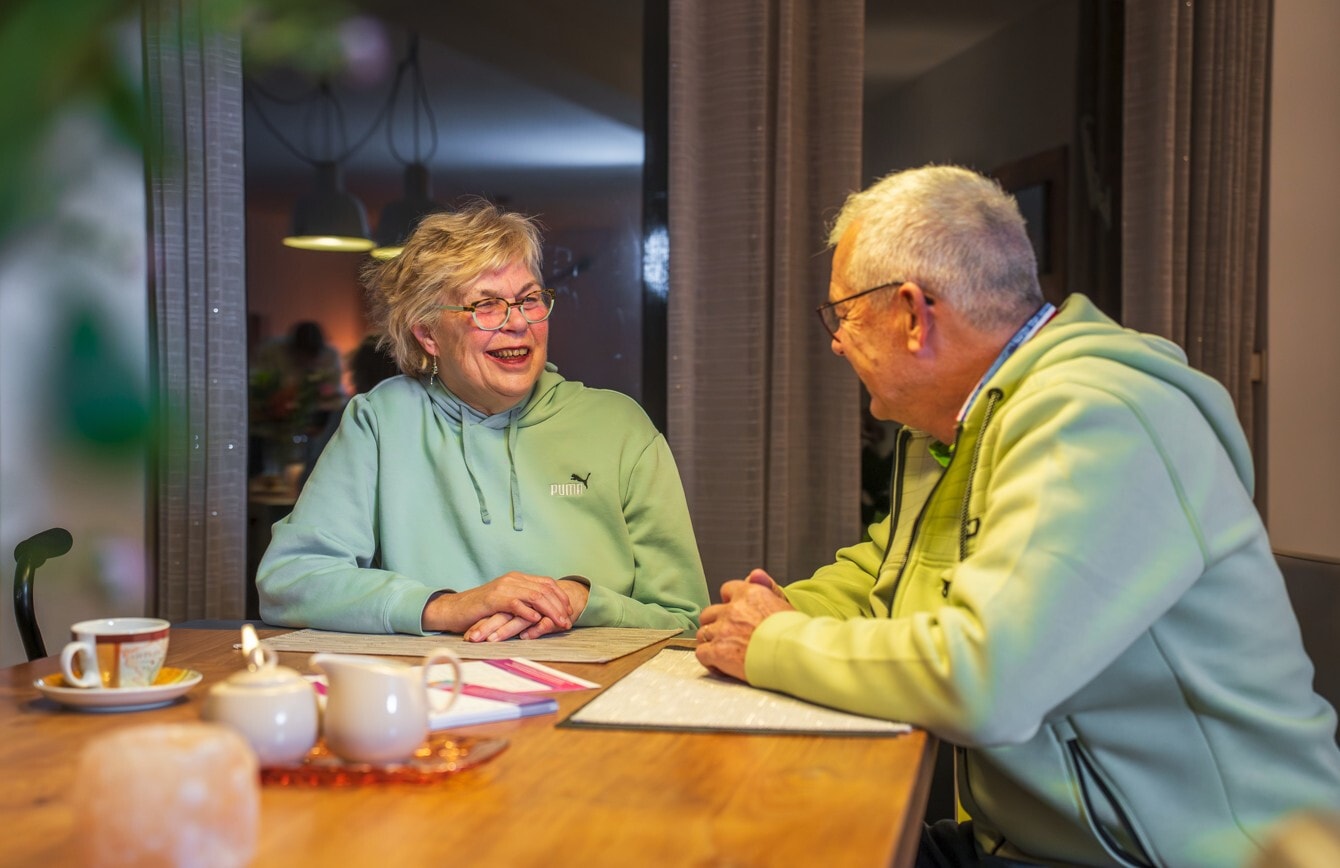 Een glimlachende oudere vrouw en een oudere man zitten tegenover elkaar aan een houten tafel en voeren een gesprek.