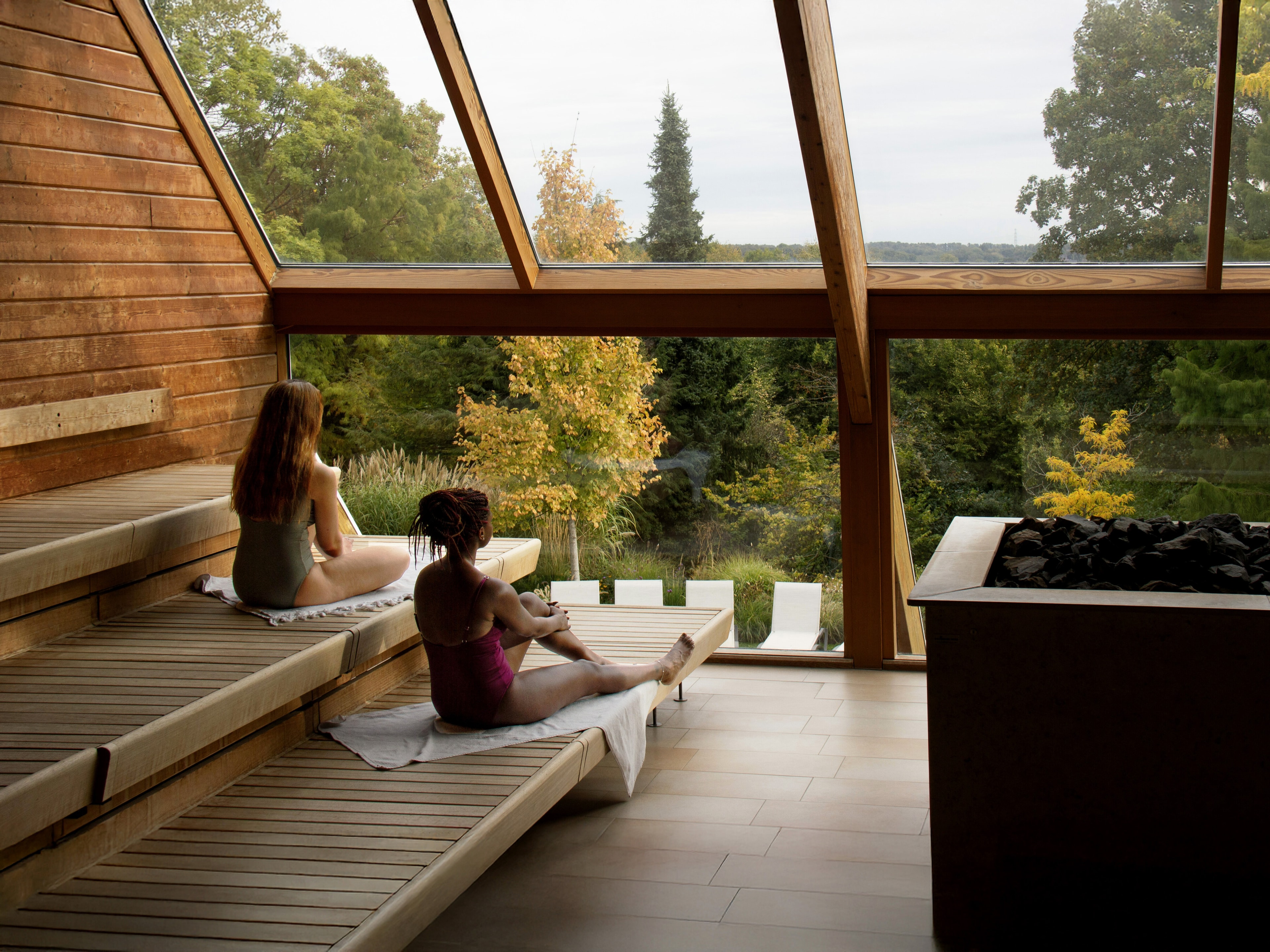 Twee vrouwen ontspannen in een panoramische houten sauna met uitzicht op een groen bos.
