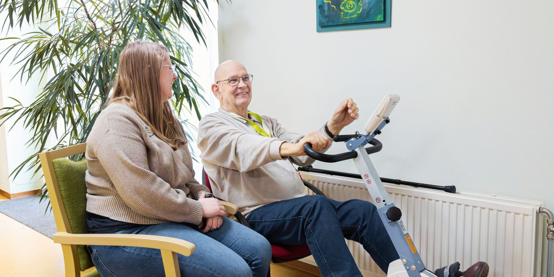 Een oudere man maakt gebruik van een fietstrainer terwijl hij een gezellig praatje maakt met een jonge vrouw in een lichte kamer met planten.
