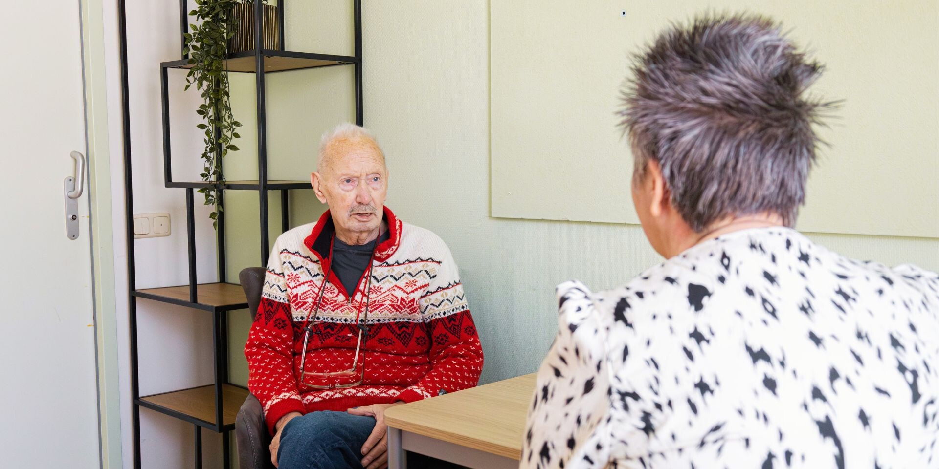 Een oudere man in een rode trui voert aan tafel een serieus gesprek met een gesprekspartner, die met haar rug naar de camera zit.