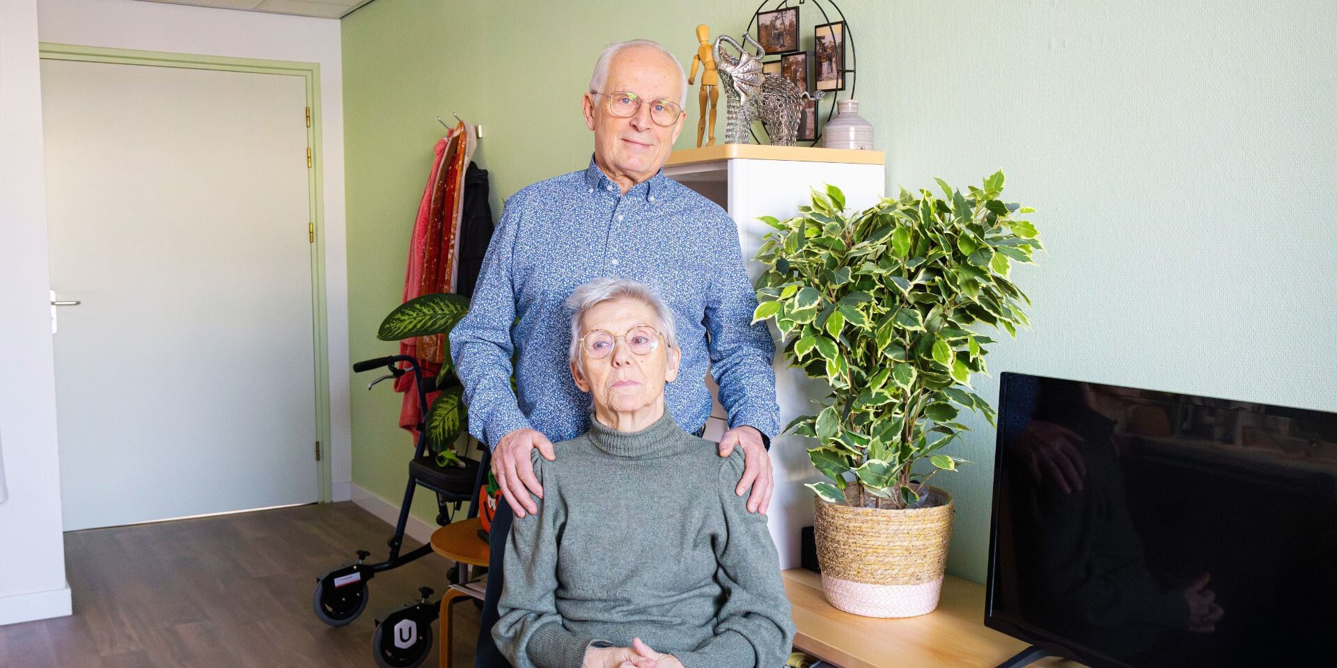 Een ouder echtpaar poseert samen in een huiselijke woonkamer; de man staat achter de zittende vrouw met zijn handen op haar schouders.